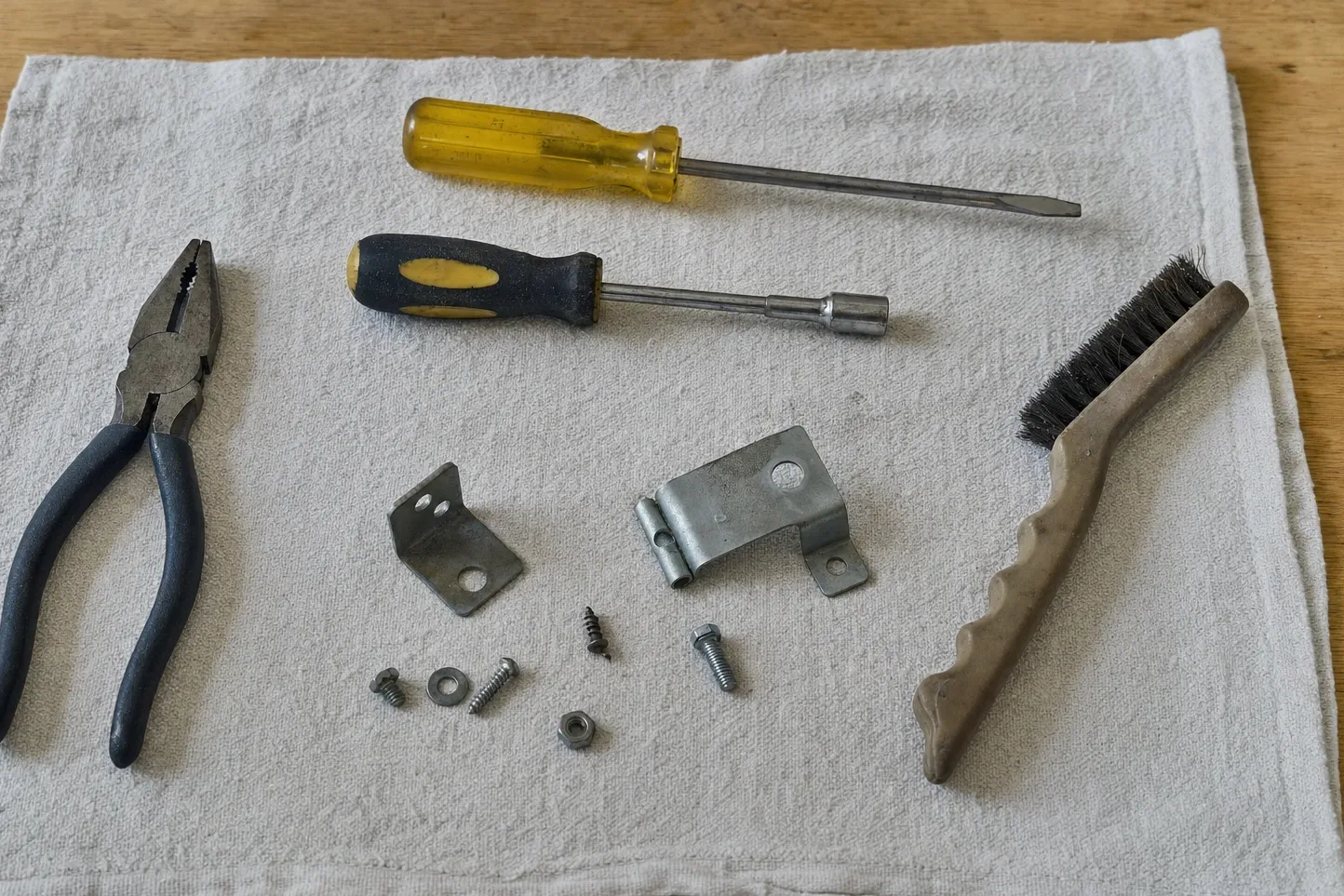 Combination pliers, a flat screwdriver, a nut driver and a wire brush with two steel brackets and loose screws on grey cloth