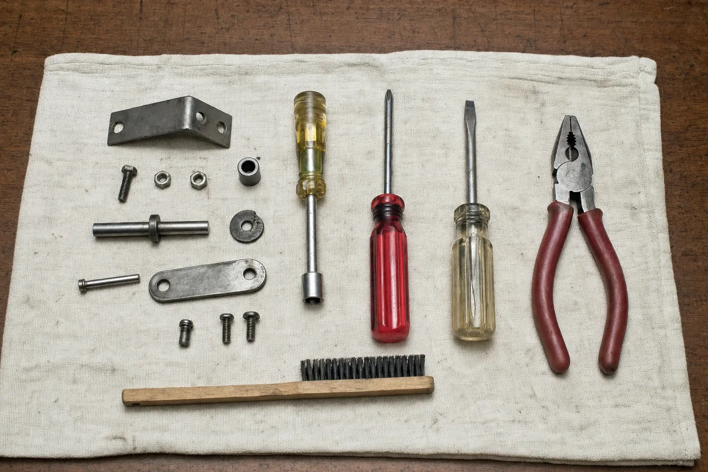 Angle bracket, shoulder pin, link strap and a bushing arranged on canvas beside a nut driver, two screwdrivers and pliers